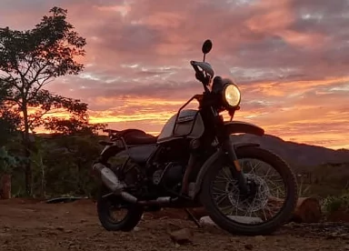 Royal Enfield Himalayan phare allumé devant un ciel de coucher de soleil orangé