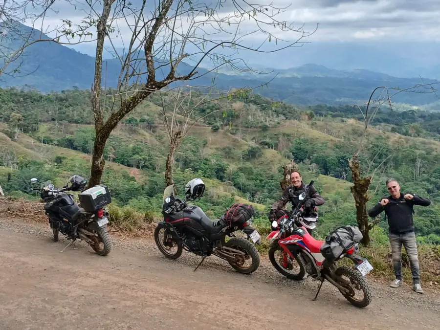 Trois motos garées au bord d'une piste et deux motards qui posent, collines en arrière-plan