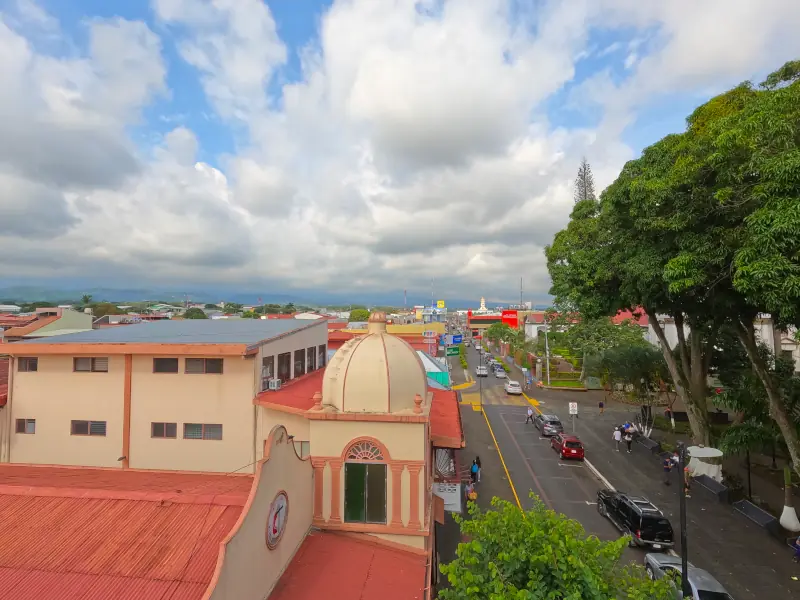 Vue en hauteur d'une rue d'Alajuela, avec un bâtiment à coupole et de grands arbres, sous un ciel nuageux