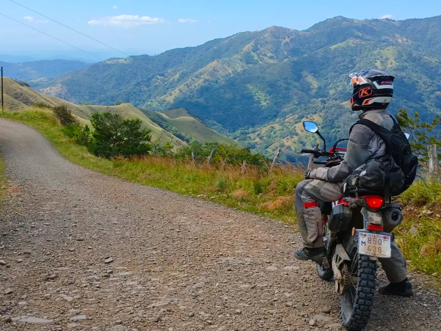 Un motard arrêté sur une piste de gravier, face aux montagnes verdoyantes